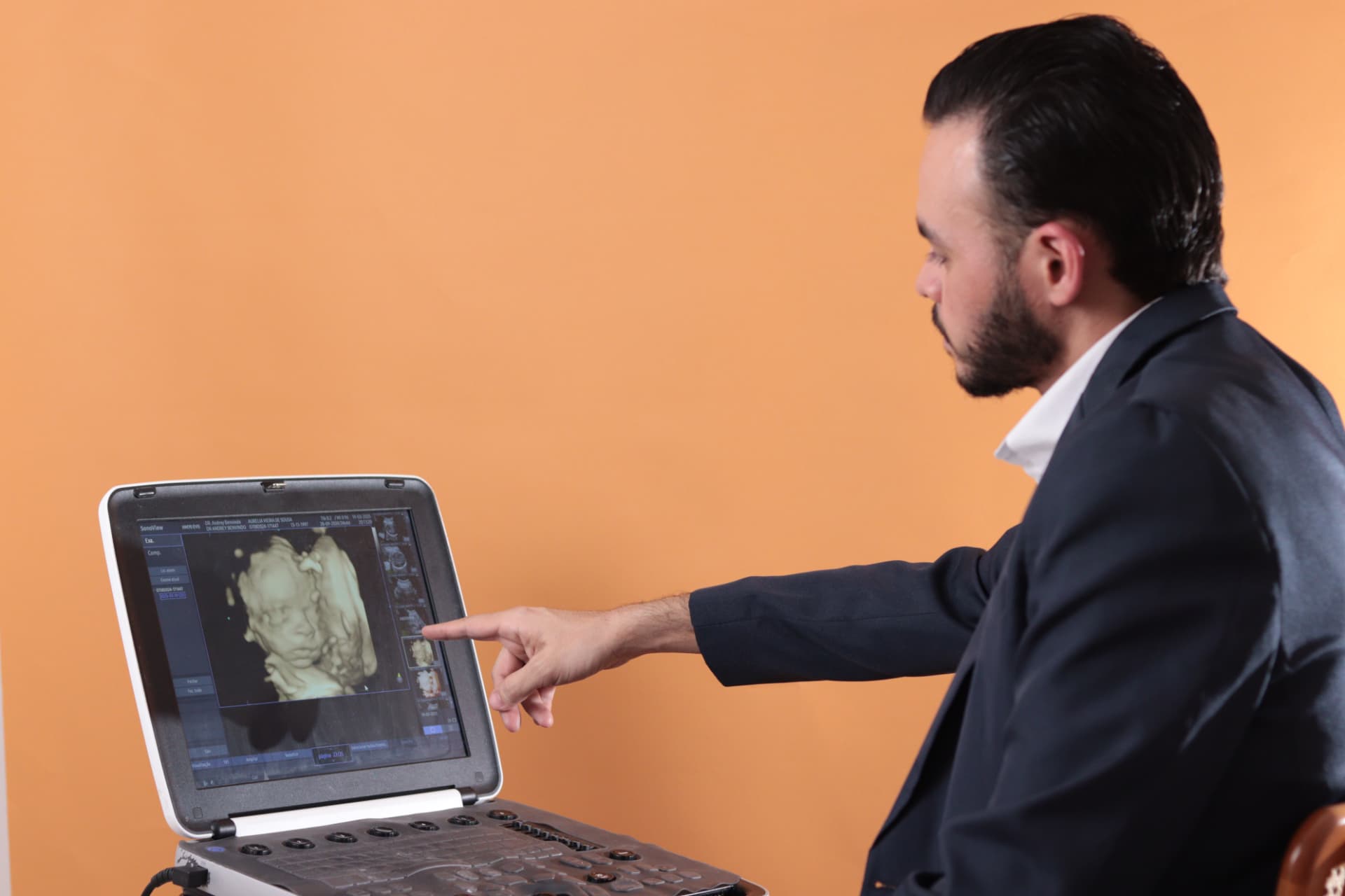 Dr. Andrey Benvindo apontando para imagem de ultrassonografia em equipamento médico com fundo laranja